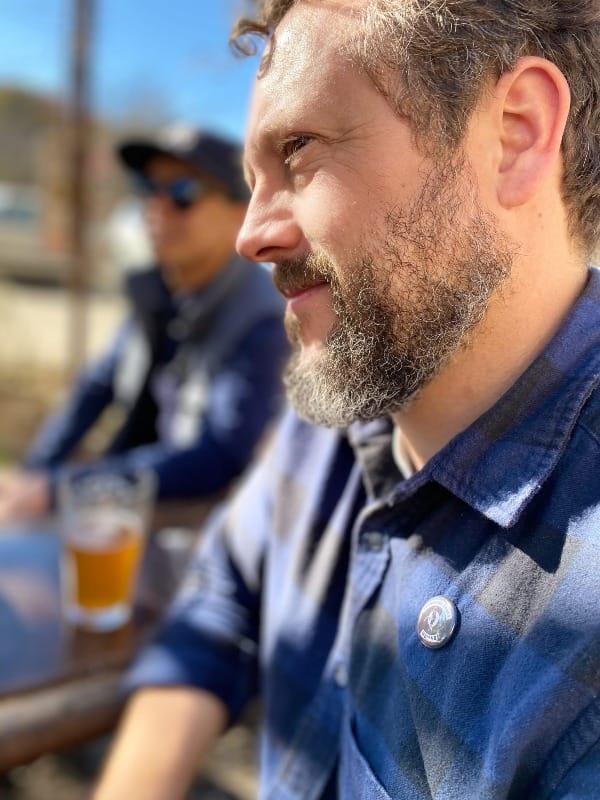 Photo of Wess Daniels from left side. Wearing a blue checkered flannel, outside on a picnic table.